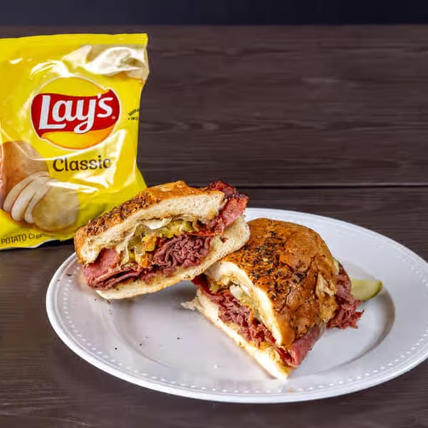 A Weinberger's Deli pastrami sandwich, cut in half, on a white plate with a bag of Lay's chips.