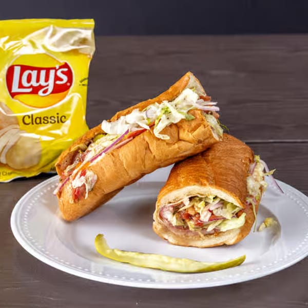 Two deli sandwiches with lettuce, tomato, and onion, a pickle spear, and a bag of Lay's chips.